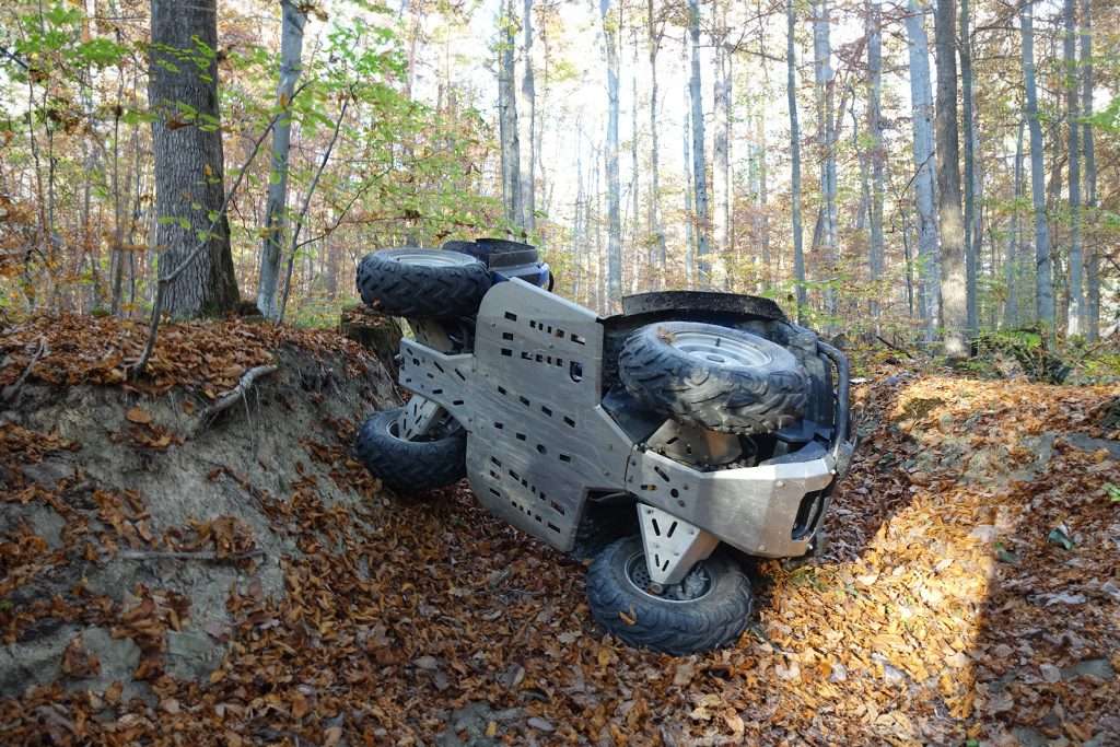 An overturned ATV lying on its side amidst a forest floor covered with autumn leaves, suggesting a recent accident or mishap during off-road driving.