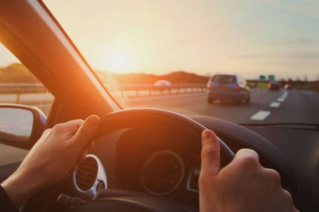A person driving a car on a highway during sunset, with their hands on the steering wheel and the warm glow of the sun ahead, carefully avoiding potential car accidents.